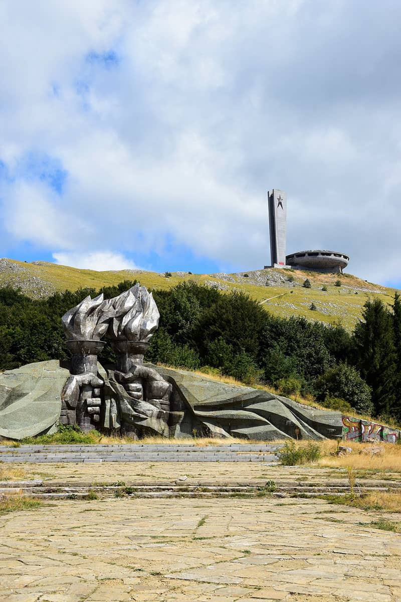 Monument approach showing socialist hands sculpture and Buzludzha UFO building on mountain peak